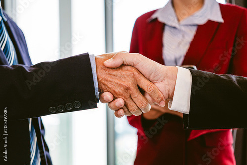 Two business professionals shake hands in a formal office setting as a colleague looks on, symbolizing a successful deal, partnership, and mutual trust
