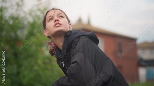Teen leaning on railing in rain, young observer scanning skyline black jacket hood damp with drizzle, redbrick buildings and leafy backdrop, thoughtful expression, cinematic urban atmosphere.