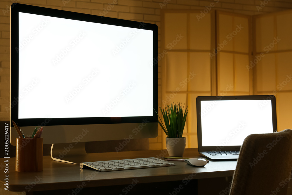 Fototapeta premium Modern programmer's workplace with computer monitor, laptop and houseplant in office at night, closeup