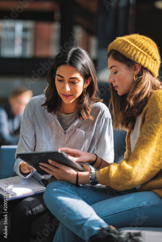 Gen z female students reviewing homework together using tablet in university lounge, medium close-up academic teamwork scene, clean campus interior, natural daylight