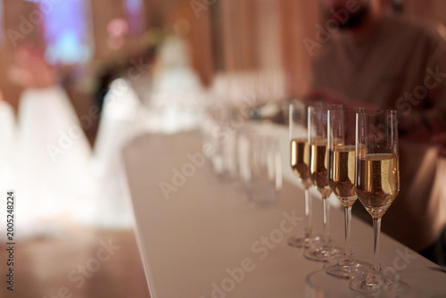 Champagne Glasses in a Row at Luxury Wedding Reception. Close-up of sparkling champagne flutes lined up on a white bar counter during a gala event or wedding. Blurred festive background