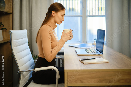 Photography Woman sits at desk holding cup while looking at laptop in home office during the