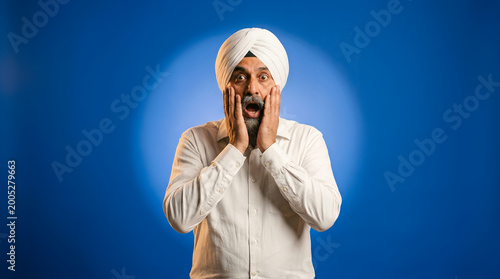 Man wearing a white turban and shirt looks shocked with hands on face