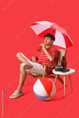 Young man with beer and beach accessories resting in chair on red background