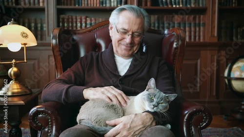Serene moments unfold as an older gentleman shares quiet joy with his affectionate cat, nestled deeply in a grand library armchair