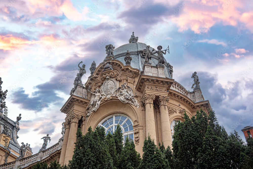 Fototapeta premium The ornate Baroque facade of Vajdahunyad Castle in Budapest features grand statues and a prominent dome set against a vibrant pink sunset sky.