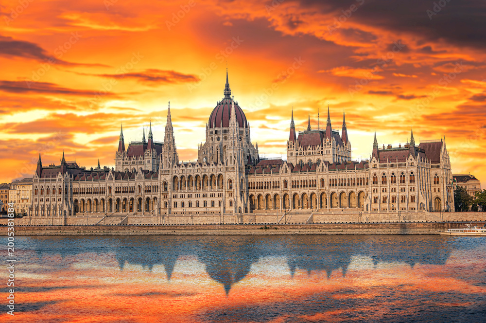 Naklejka premium The Hungarian Parliament Building stands majestically along the Danube River under a vibrant orange sunset sky.
