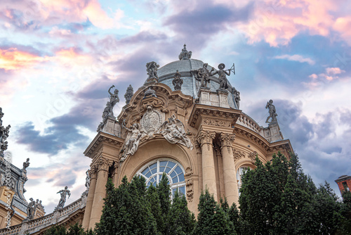 The ornate Baroque facade of Vajdahunyad Castle in Budapest features grand statues and a prominent dome set against a vibrant pink sunset sky.