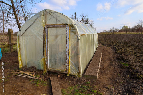 A homemade greenhouse with a wooden frame covered in thick plastic wrap stands on a prepared garden plot in early spring. Nearby is a long wooden garden bed with loose soil under an open blue sky.