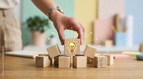 Creative professional hand selects a wooden block featuring a glowing light bulb icon to signify the arrival of a brilliant idea within a collaborative work environment.