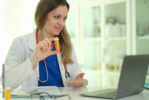 Doctor holding prescription bottle during telehealth consultation via laptop, counseling patient on dosage, side effects and safe