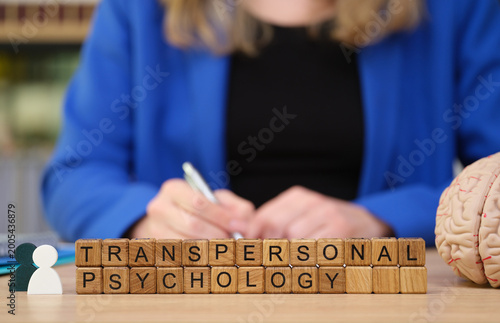 On a study table, a person in a blue jacket writes notes, focused on transpersonal psychology. Wooden blocks spell out the subject beside a model brain, capturing the essence of mental exploration.