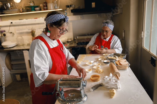 Two elderly female chefs wearing red aprons and headbands prepare handmade dough and tarts in a bright commercial bakery kitchen