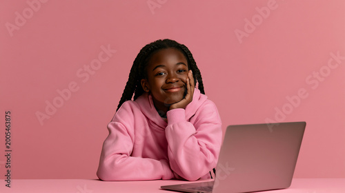 cheerful african teenage girl using laptop at pink background, smiling young student working on laptop at modern table, happy adolescent girl studying online with computer indoors