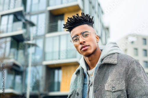 A young man with glasses and styled hair stands confidently in front of a modern apartment building.