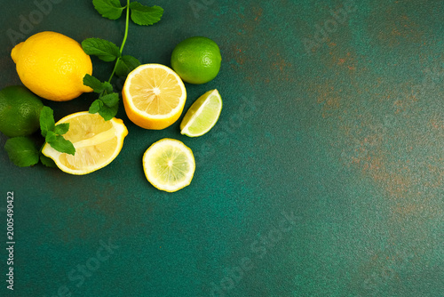 Fotografija Fresh lemons and limes sliced with green leaves on dark green table surface in b