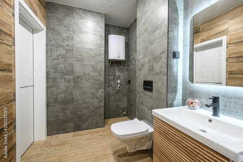 modern bathroom with grey textured walls, wooden floor and vanity, wall-mounted toilet, sleek black faucet, illuminated mirror, white sink, and water heater mounted on the wall.