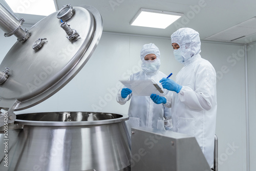 Two technicians in protective suits check mixing equipment in. Professional scientists in cleanroom suits inspecting pharmaceutical manufacturing processes and operating industrial mixing equipment.