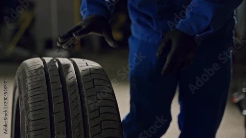 Mechanic in blue coveralls wearing black gloves rolling a car tire with visible tread pattern inside an auto repair workshop, demonstrating maintenance and inspection of the wheel