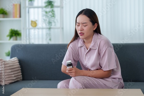 Asian women suffering stomach abdominal pain holding belly medicine pill bottle sitting couch sofa wearing pink lavender pajamas healthcare wellness pain relief home