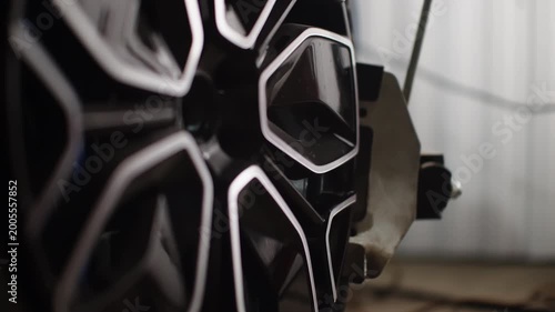 Macro close-up of a glossy black alloy wheel with intricate geometric spokes beside a mounted brake caliper in an automotive workshop, highlighting precision machining and finishes