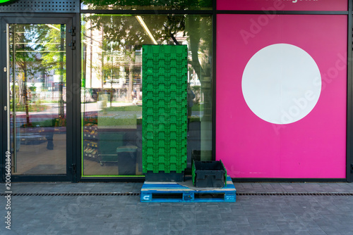 Stacked green plastic produce crates stand on a pallet outside a glass grocery storefront with a bright pink wall panel and large white circle. Useful for retail supply, food store logistics and urban