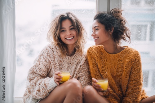 Two Happy Friends Enjoying Fresh Orange Juice by Window on Cozy Winter Day