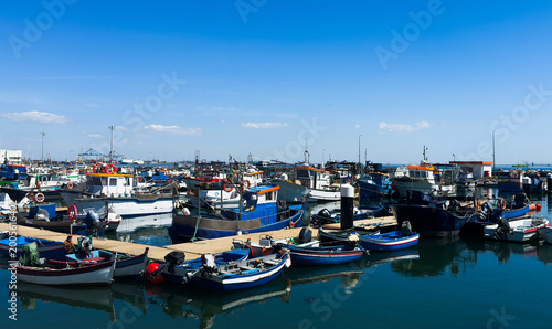 Colorful traditional fishing boats moored in the calm, reflective water of Setubal harbor, Portugal. Stone steps and a beached boat on the shore. Setubal, Portugal. 