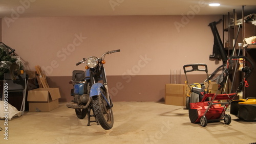 Quadro su tela Old blue motorcycle resting on its kickstand in a dimly lit, cluttered garage or