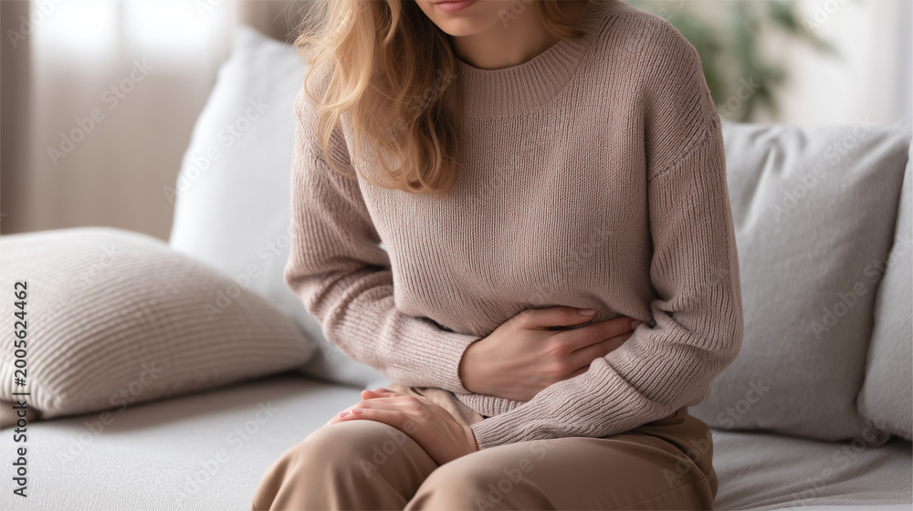 Fototapeta premium Young woman sitting on sofa holding stomach in pain, abdominal discomfort and digestive issue, healthcare and lifestyle concept.