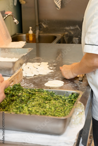 In a semi-open kitchen, chefs carefully prepare xiaolongbao by flattening dough into thin wrappers and filling them with minced pork and chives. Their hands move with precision, folding delicate pleat