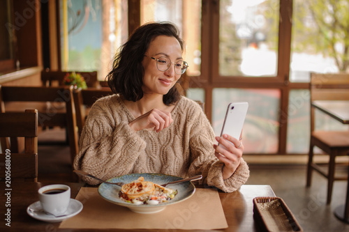 Positive asian woman in eyeglasses using mobile phone near salad and coffee in cafe