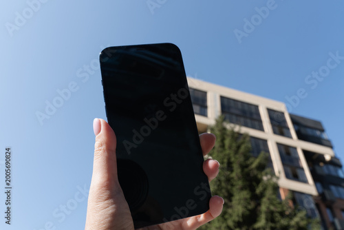 Woman’s hand holding a smartphone outdoors with a modern building and green tree in the background, blending digital and urban nature concepts.