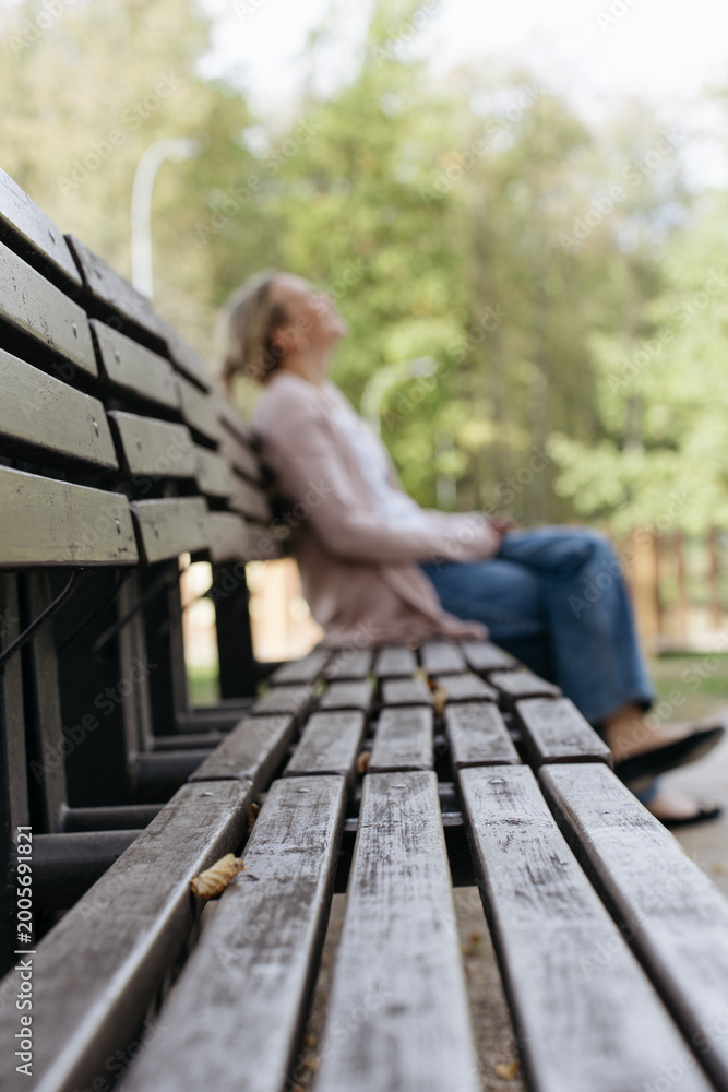 Fototapeta premium Selective focus view of a wooden park bench with a blurred caucasian woman resting in the background, ideal for solitude, reflection and mental wellness concepts.