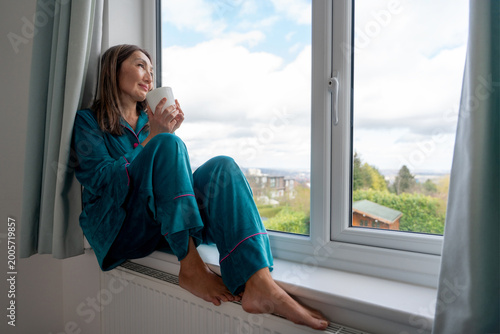 Free Download Woman sits on the window sill with a cup, looking outside at the cloudy sky and greenery in the early morning   - GFXLoad