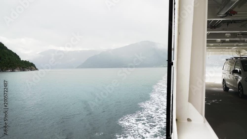 View from a Norwegian ferry crossing a scenic fjord with a car parked on the open deck. Misty mountain landscape and water ripples in Norway. Travel and nordic transportation. 4K