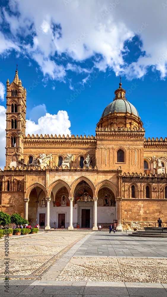 Fototapeta premium Large ornate cathedral with detailed architecture under a bright blue sky filled with fluffy white clouds, with arched walkways
