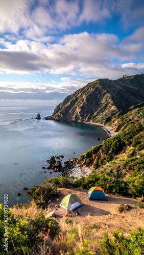 Scenic coastal campsite with tents set up, surrounded by mountains and sea, under a cloudy, blue sky