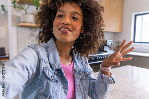 African American woman filming selfie at kitchen counter, wearing denim jacket, hoops and rings