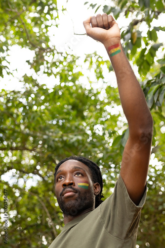 Man raising fist skyward under leafy canopy wearing olive shirt bearing Pan-African stripes