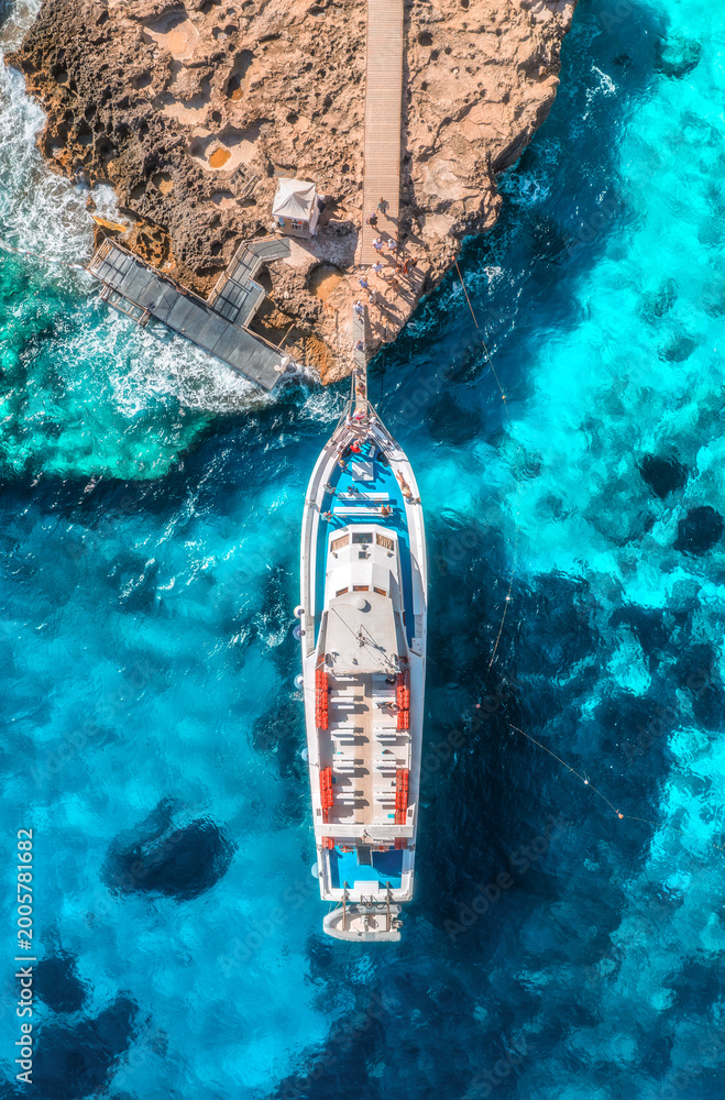 Fototapeta premium Aerial view of boat with people and rocky sea coast on sunny summer day. Top drone view of yacht, transparent azure water, path. Mediterranean sea. Travel in Sardinia island, Italy. Tropical landscape