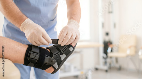 A medical professional applies a support brace to a patient's injured ankle. The setting is a clinic with equipment in the background. The staff wears gloves for safety