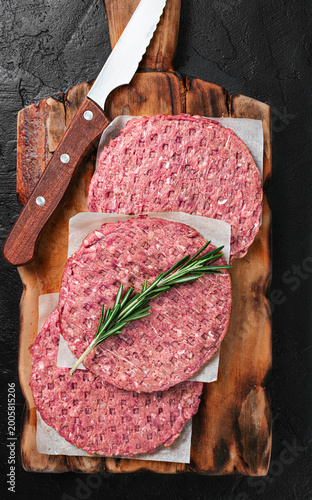 Raw beef burger patties with fresh rosemary and a kitchen knife on a wooden cutting board, dark stone background, top view.