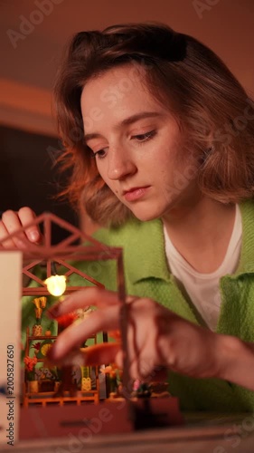 Young woman meticulously assembling a detailed miniature DIY greenhouse kit by hand. Focus on her hands and the tiny intricate plants. Creative hobby, mindfulness, and craftsmanship concept photo.