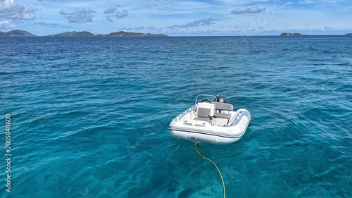 Fényképezés White inflatable dinghy floats on clear turquoise Caribbean water, tethered by rope, with distant islands and soft clouds under blue sky, creating a calm, minimal ocean scene