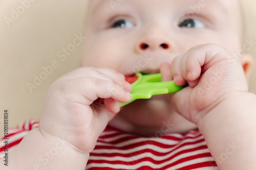 Baby lying on bed and gripping teething ring with both hands. Child chewing toy for teething relief. Exploring textures while soothing sore gums. Sensory discovery at home.
