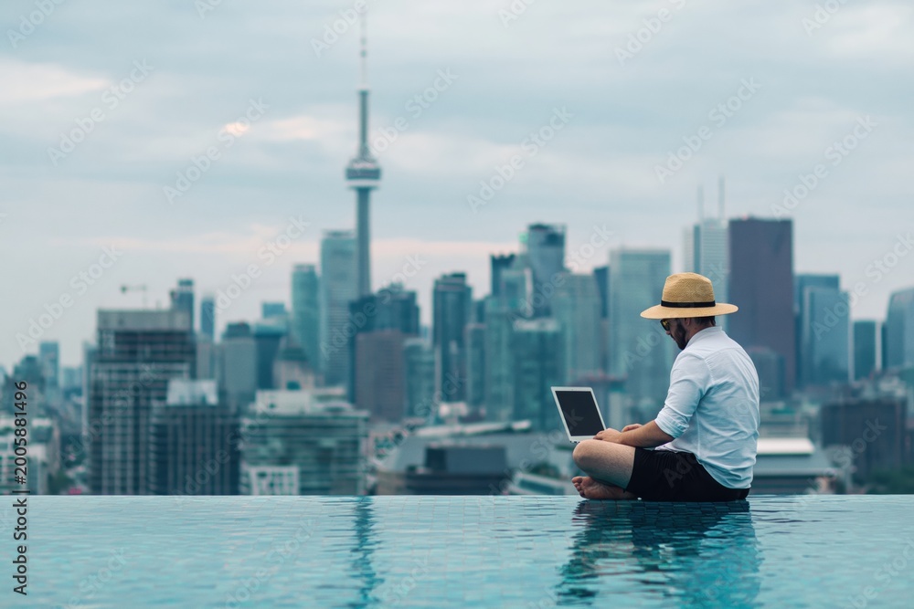 Fototapeta premium Man in a straw hat using a laptop by an infinity pool, overlooking the Toronto city skyline with the CN Tower