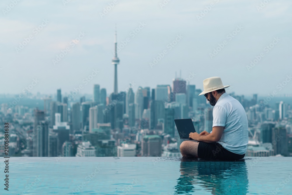 Fototapeta premium Man using laptop by infinity pool overlooking Toronto skyline and CN Tower. Digital nomad working remotely with freedom to travel
