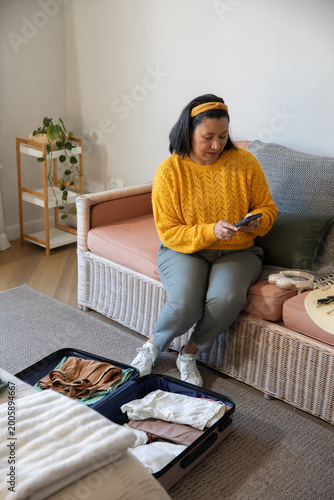 Mature woman wearing yellow sweater sitting on sofa at home packing suitcase while using smartphone