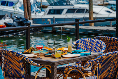 Outdoor dining on a marina with food and drinks near boats. People enjoy food and drinks at a table by water with boats in the background.